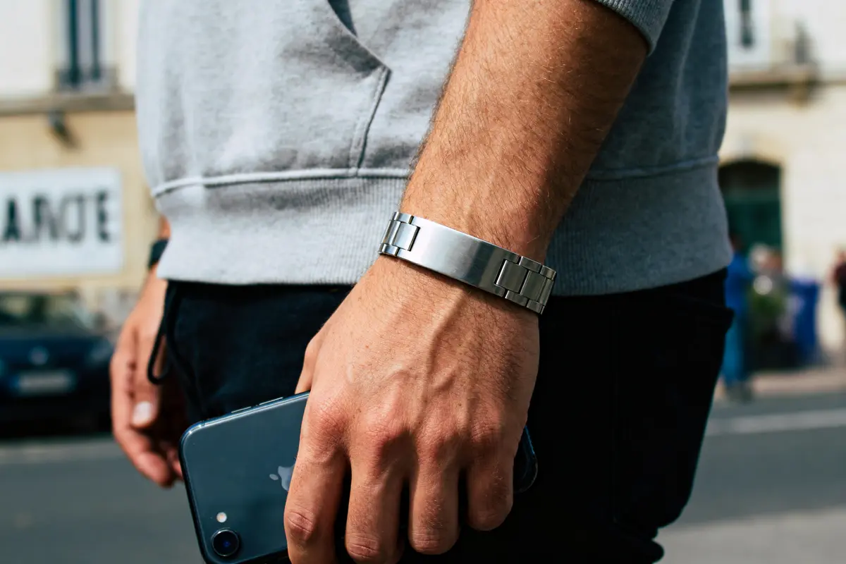Homme tenant un smartphone, bracelet métallique au poignet, portant un sweat gris, en extérieur urbain flou.
