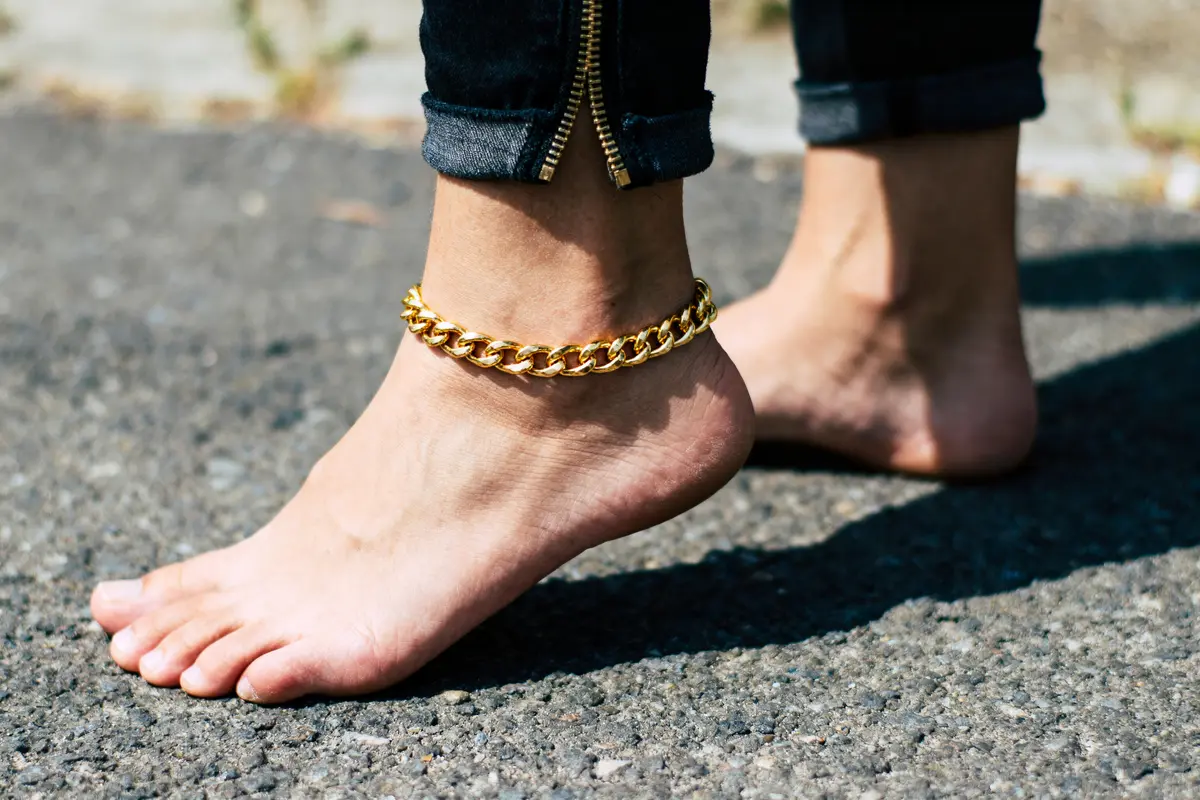 Personne pieds nus portant un bracelet de cheville métallique doré, debout sur une route asphaltée.