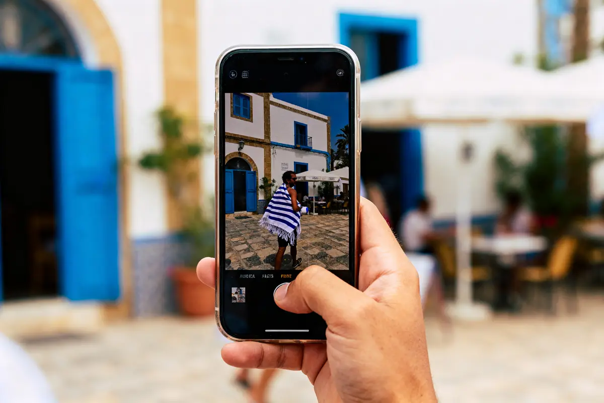 Main tenant un smartphone devant bâtiment blanc et bleu, avec personne enroulée dans une serviette rayée.