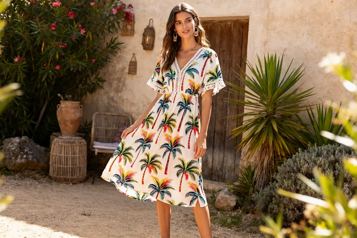 Une femme porte une robe à motifs de palmiers devant une maison méditerranéenne avec plantes.