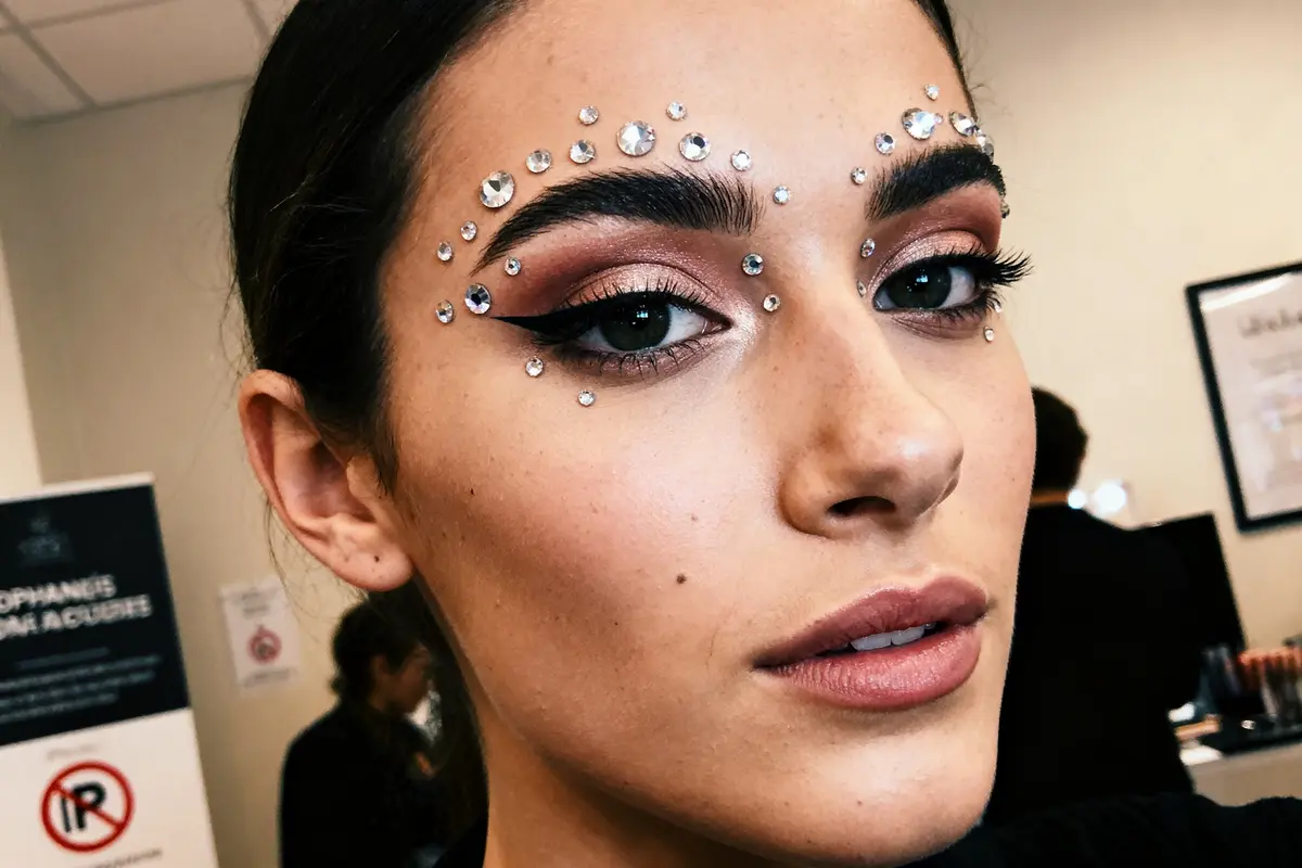 Femme avec maquillage festif, pierres scintillantes autour des yeux, yeux soulignés, ombre à paupières cuivrée.